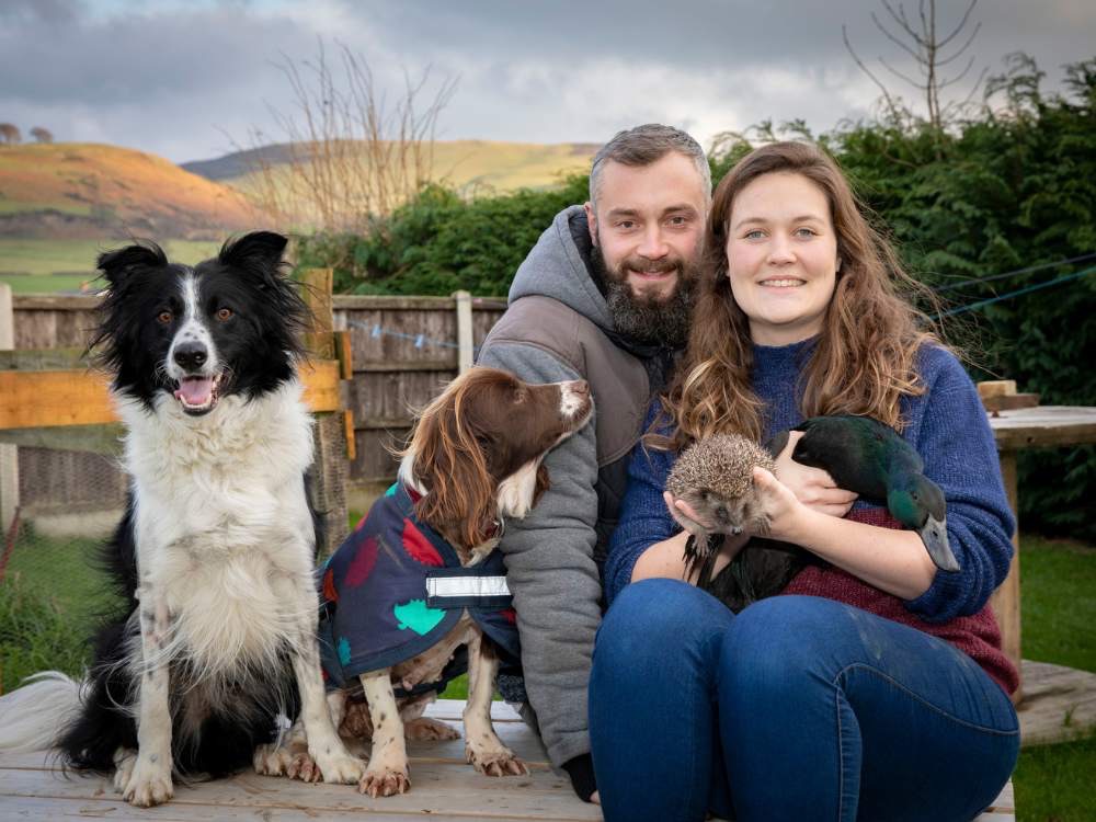 Young couple sitting outside with two dogs, a hedgehog and a duck