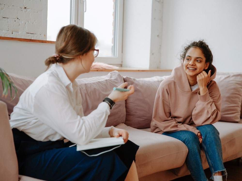 Young woman in pink hooded top sitting on sofa smiling and talking to counsellor