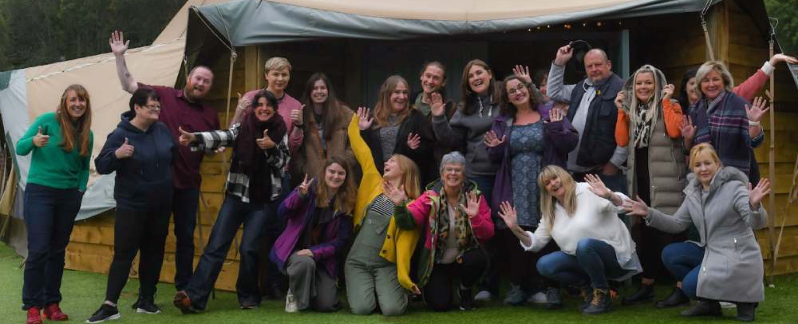 Group of twenty adults kneeling and standing outside in front of a tent all waving to the camera smiling