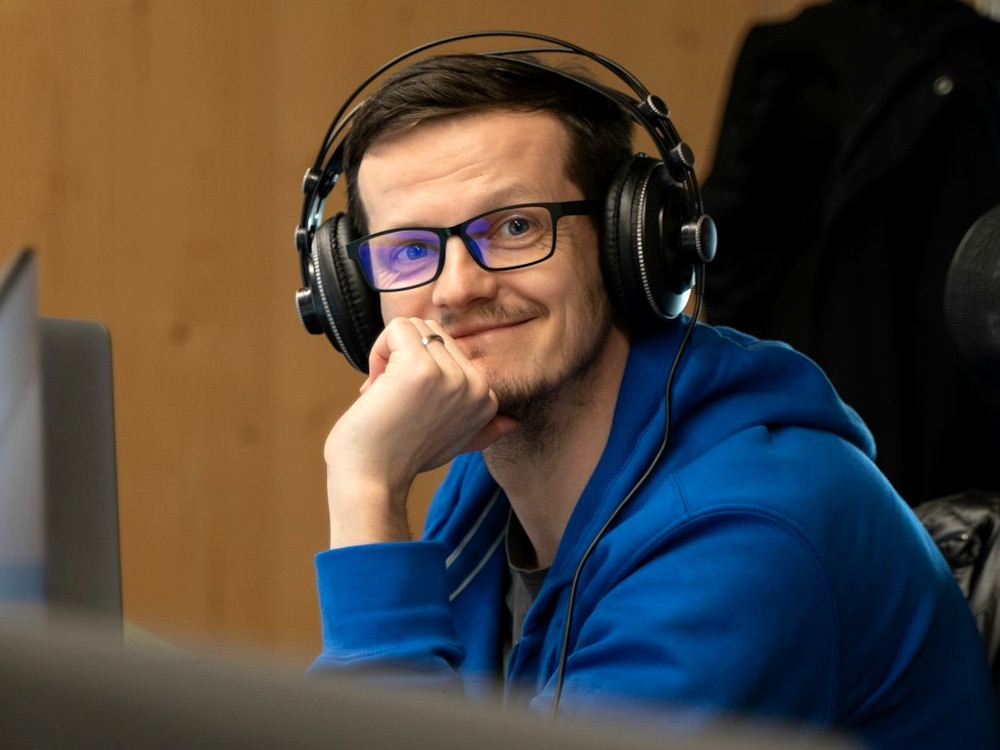 Young male with glasses and blue hooded top looking at the camera and smiling in work